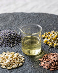 A small clear glass beaker filled with pale yellow liquid sits on a dark stone slab, surrounded by small piles of dried botanical ingredients including lavender, chamomile flowers, jasmine, and cedarwood pieces.