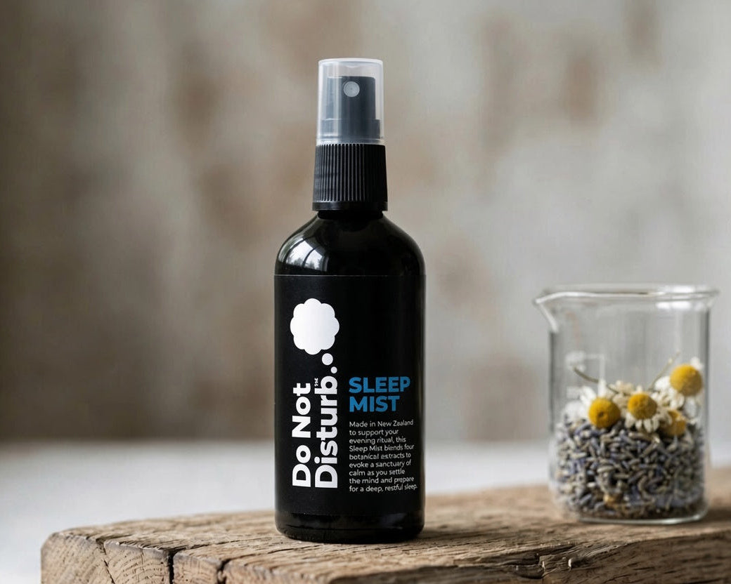 A black spray bottle labeled 'Do Not Disturb Sleep Mist' stands on a rustic wooden surface next to a small glass beaker containing dried lavender and chamomile flowers, with a softly blurred neutral background.