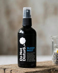 A black spray bottle labeled 'Do Not Disturb Sleep Mist' stands on a rustic wooden surface next to a small glass beaker containing dried lavender and chamomile flowers, with a softly blurred neutral background.