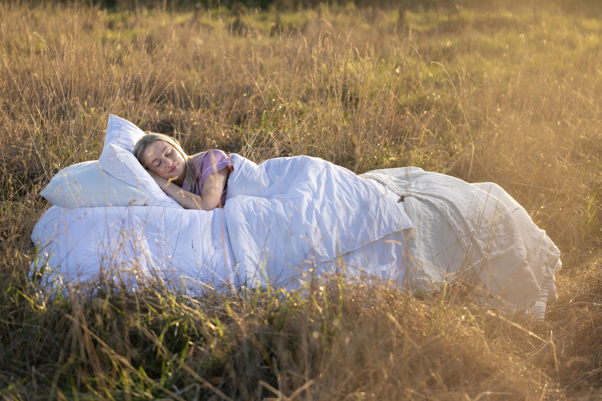 A woman lies on her back on a bed made from a large white duvet and pillows, set outdoors in a field of dry golden grass at sunset.