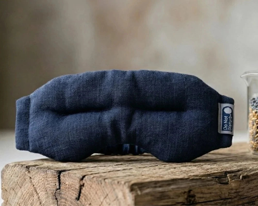 Weighted eye mask in Midnight Blue on a wooden block, with a jar of flaxseed & lavender beside it